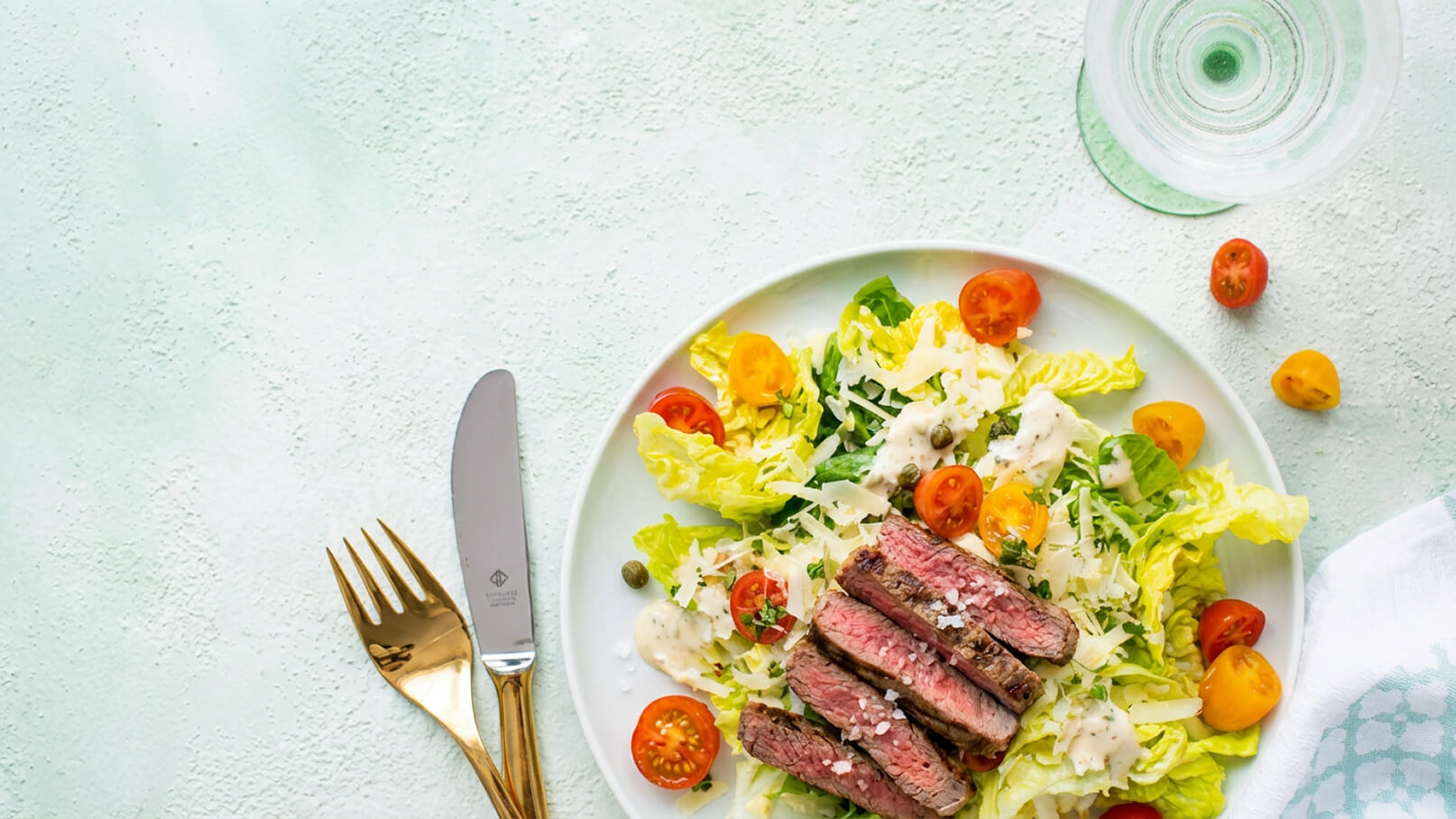 Ein Teller mit frischem Salat, grünem Blattgemüse, Kirschtomaten und cremigem Dressing. Darauf liegen in Scheiben geschnittenes Steak mit einer knusprigen Kruste. Neben dem Teller liegen eine goldene Gabel und ein Messer.