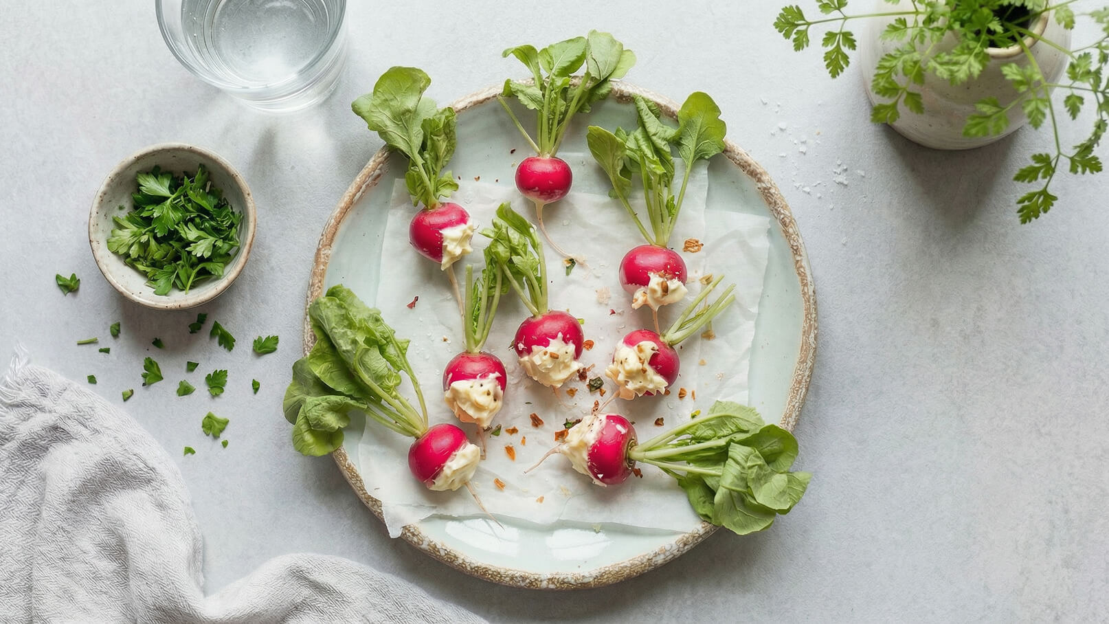 Ein Teller mit frischen, rot-weißen Radieschen, gefüllt mit einer cremigen Füllung und umgeben von grünen Blättern. Auf dem Tisch liegen gehackte Kräuter und eine Schale mit frischer Petersilie. Im Hintergrund steht ein Glas Wasser.