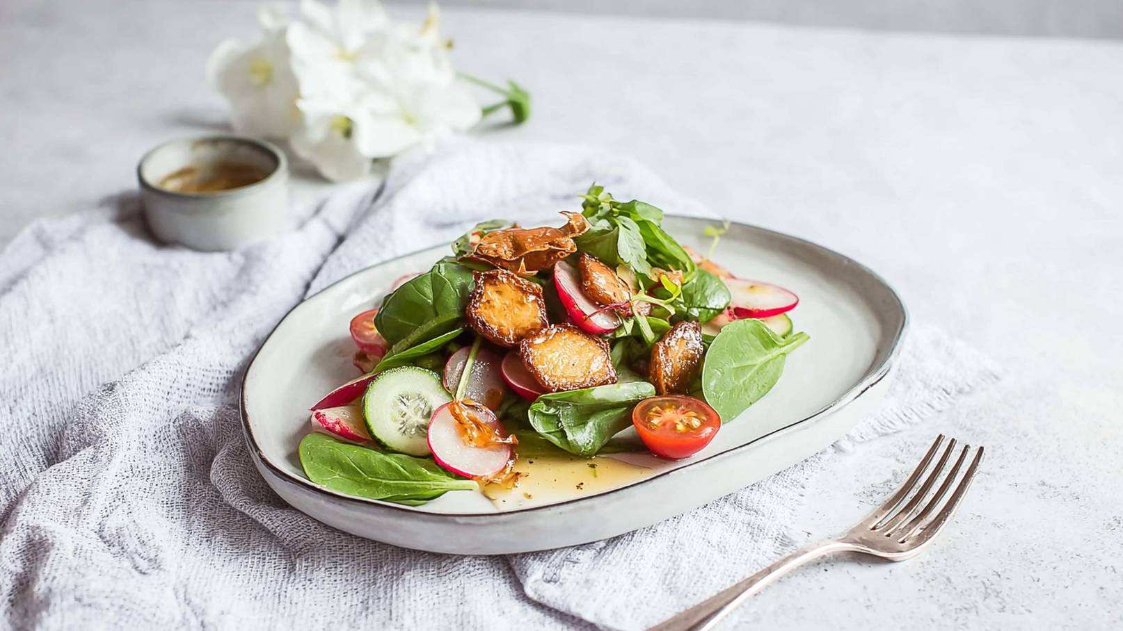 Ein frischer Salat auf einem eleganten Tellerrand. Die Zutaten umfassen Spinat, Radieschen, Gurken und Kirschtomaten, garniert mit knusprigen Süßkartoffeln. Daneben steht eine kleine Schüssel mit Dressing. Der Hintergrund ist hell und neutral.