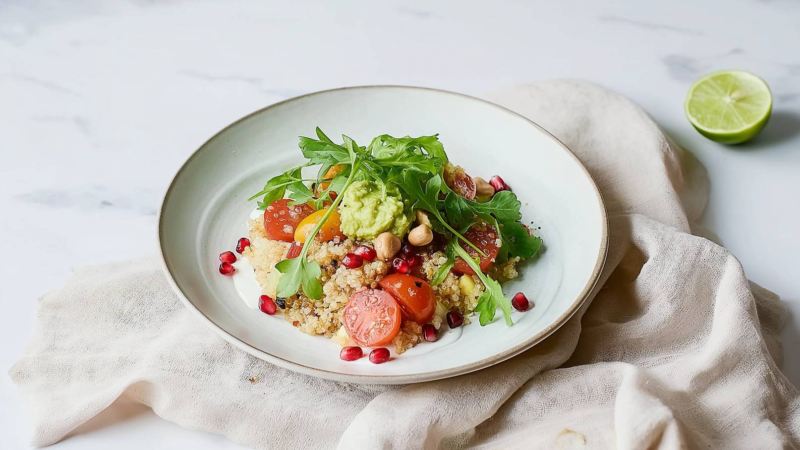 Eine Schüssel mit Quinoa, garniert mit Kirschtomaten, Granatapfelkernen, Erdnüssen, Avocadocreme und frischem Rucola. Die Farben sind lebhaft und appetitlich, mit einem Hauch von Limette im Hintergrund.