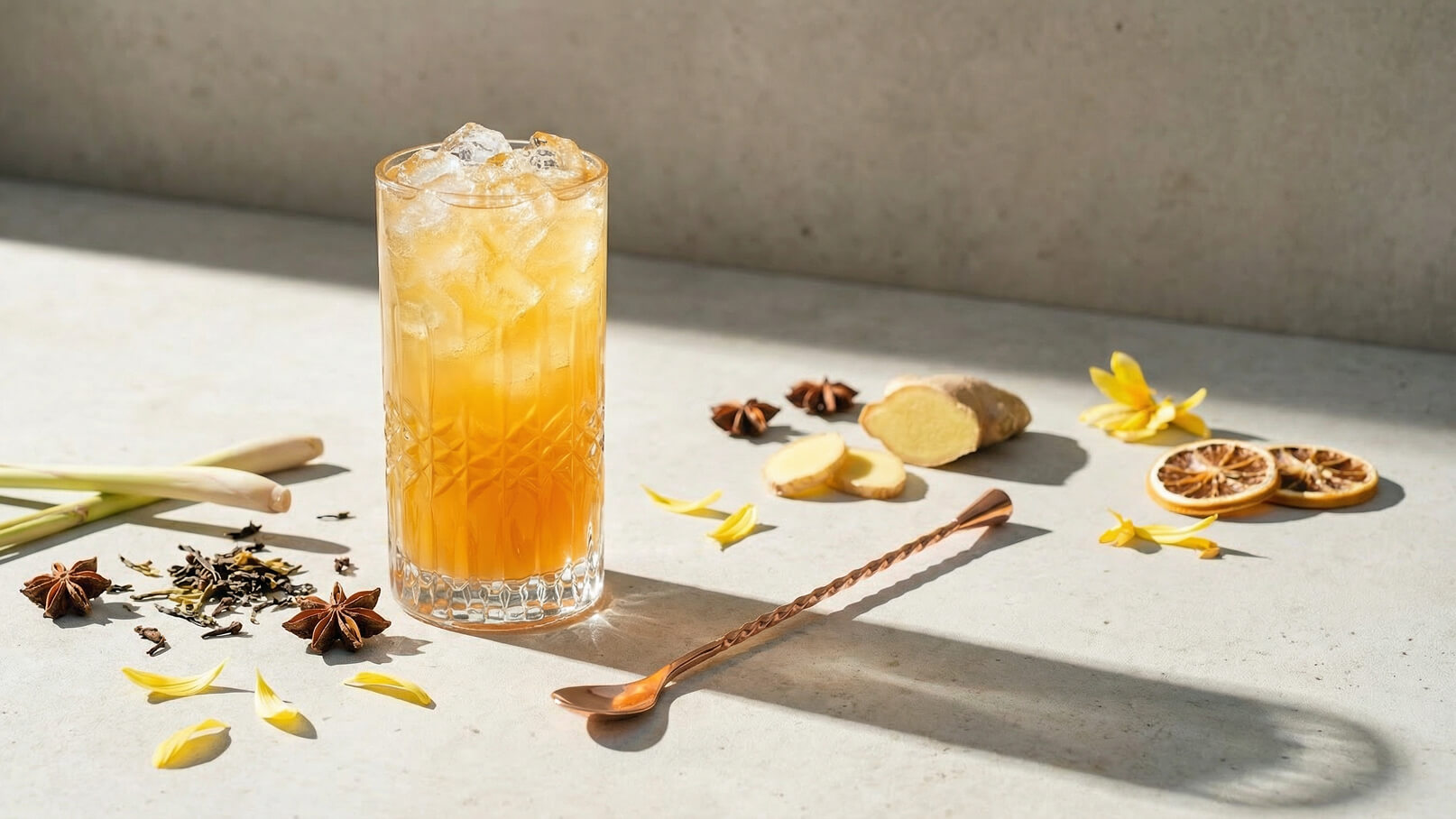 Ein erfrischendes Getränk in einem hohen Glas, gefüllt mit Eiswürfeln und einer orangefarbenen Flüssigkeit. Rund um das Glas sind aromatische Zutaten wie Ingwer, getrocknete Zitrone, Sternanis, Zitronengras und Blütenblätter verteilt, die eine sommerliche Atmosphäre vermitteln.