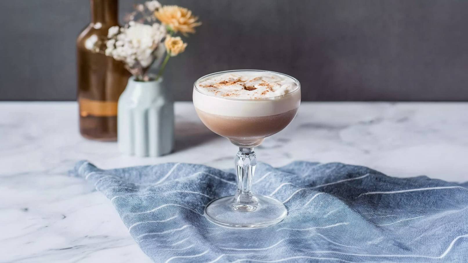 Ein elegantes Cocktailglas steht auf einem Tisch, gefüllt mit einer schaumigen Schicht Getränk, das in hellen Brauntönen gehalten ist. Oben ist eine dekorative Schicht aus Sahne und einer Prise Zimt sichtbar. Im Hintergrund sind eine flaschenförmige Vase und trockene Blumen.