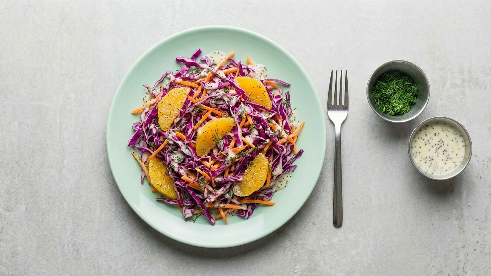 Ein Teller mit bunter Krautsalat-Mischung aus roten und orangefarbenen Zutaten, garniert mit Orangenstücken. Daneben ein Besteck und zwei kleine Schalen mit frischem Dill und einer hellen, cremigen Salatsauce.