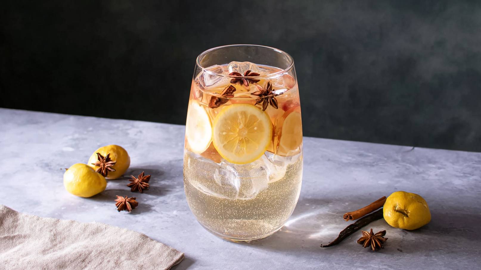 Ein Glas mit einem sprudelnden Getränk, gefüllt mit Eiswürfeln und Scheiben von Zitrone, dekoriert mit Sternanis. Auf dem Tisch liegen zwei Zitronen und einige Gewürze, darunter Zimt und weitere Sternanis. Der Hintergrund ist dunkel und schafft eine stilvolle Atmosphäre.