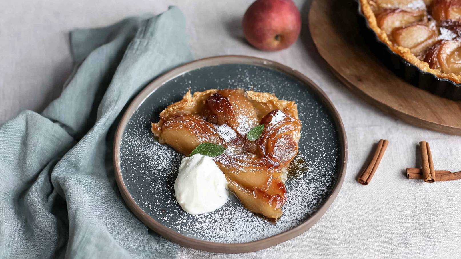 Ein Stück Apfeltarte auf einem grauen Teller, garniert mit Sahne und frischen Minzblättern. Die Tarte ist goldbraun und hat karamellisierte Äpfel, bestreut mit Puderzucker. Im Hintergrund ist ein weiteres Stück Tarte und ein roter Apfel sichtbar.