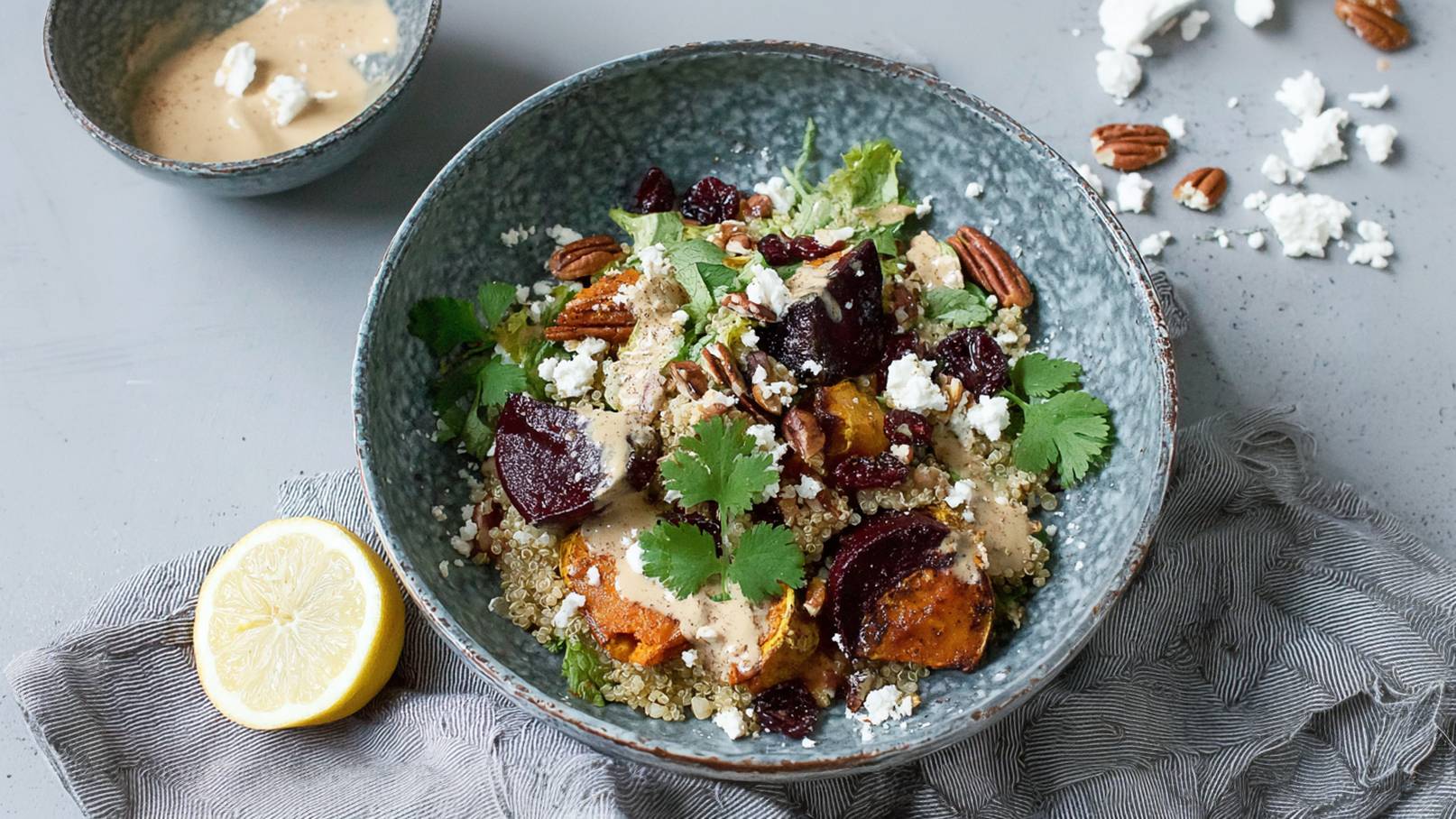 Eine Schüssel mit einem bunten Salat aus Quinoa, Rote Beete, geröstetem Gemüse, Pecanüssen und frischen Kräutern. Die Mischung ist mit einer cremigen Sauce überzogen. Neben der Schüssel liegt eine halbierte Zitrone.