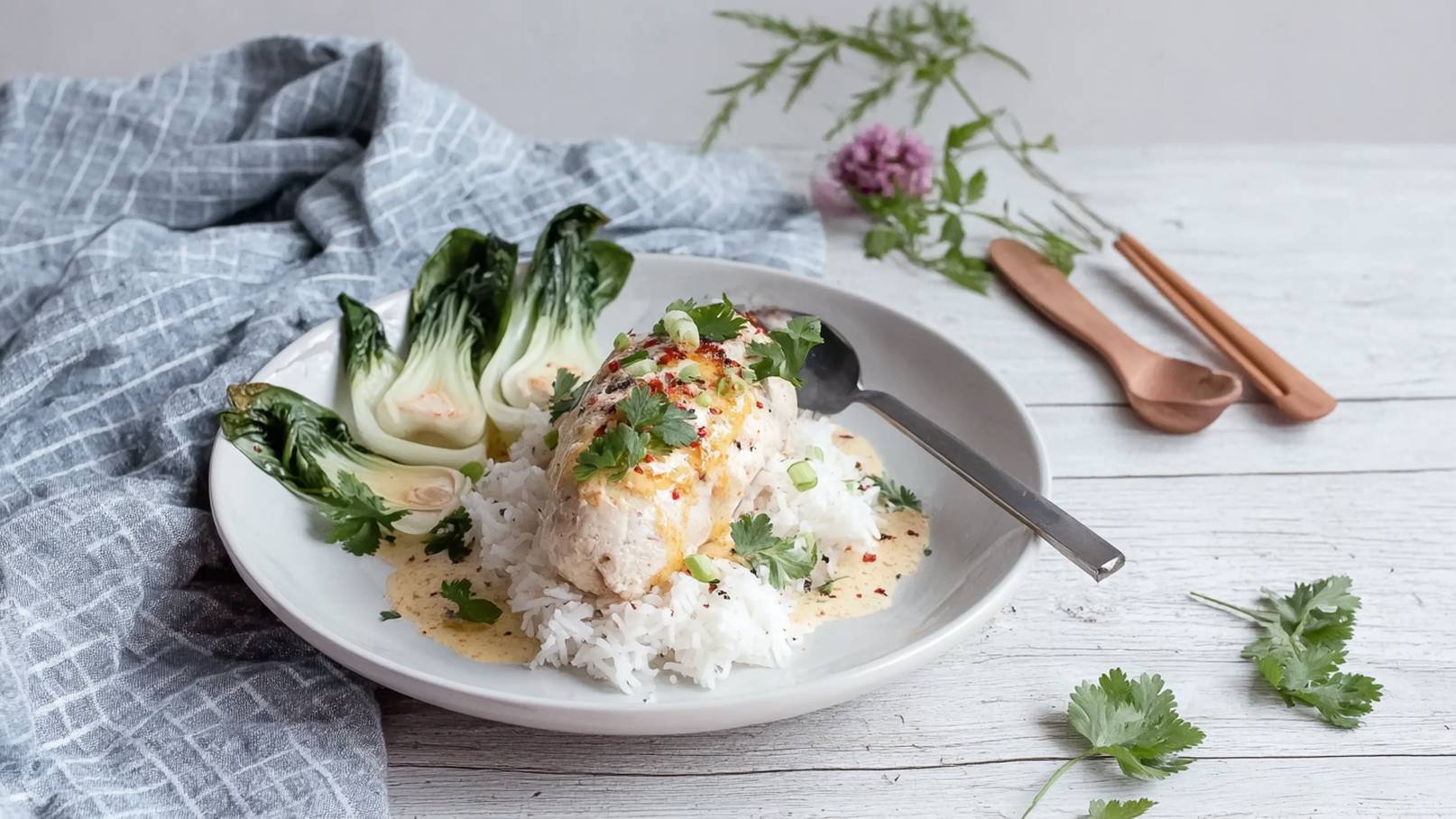 Ein Teller mit gedämpftem Reis, einem Stück Fisch in Sauce, garniert mit grünen Frühlingszwiebeln und frischem Koriander. Neben dem Fisch liegen zarte Pak Choi-Blätter. Im Hintergrund sind ein graues Tuch und Holzbesteck sichtbar.