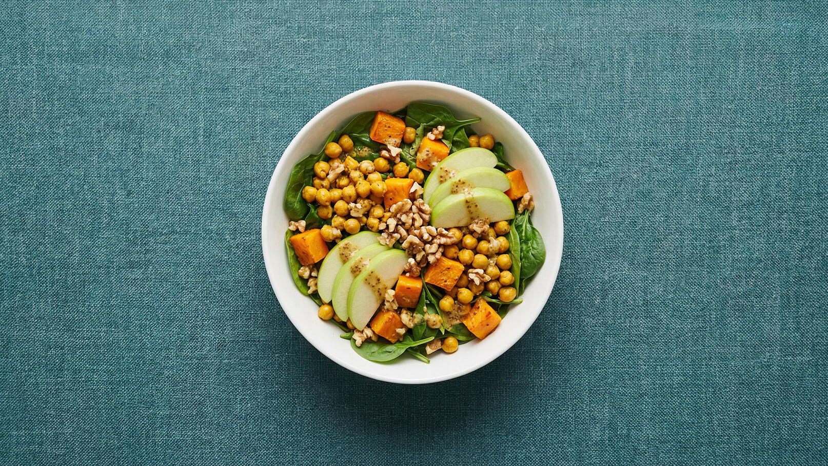 Eine Schüssel mit grünem Spinat, garniert mit Kichererbsen, Kürbiswürfeln, dünnen Apfelscheiben und Walnüssen. Die Mischung ist ansprechend und gesund, mit einer leichten Vinaigrette darüber. Der Hintergrund ist in einem sanften Blau gehalten.