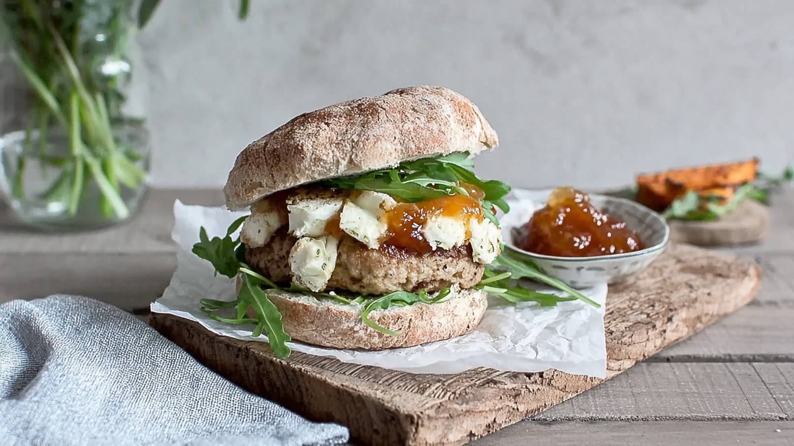 Ein frisch zubereiteter Burger auf einem Holzbrett. Der Burger besteht aus einem herzhaften Pattie, Feta-Käse, Rucola und einer süßen Sauce. Daneben befindet sich eine kleine Schale mit einer ähnlichen Sauce und etwas gebratenem Gemüse.