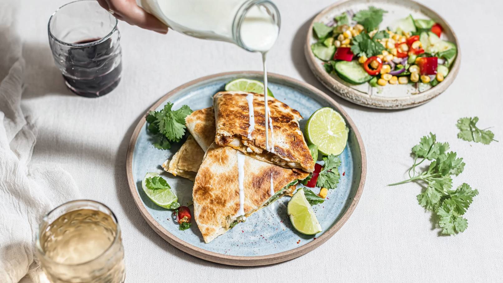 Ein Teller mit zwei knusprigen Quesadillas, garniert mit Koriander und Limettenscheiben. Sahne wird über die Quesadillas gegossen. Im Hintergrund sieht man einen bunten Salat mit Gurken, Paprika und Mais.