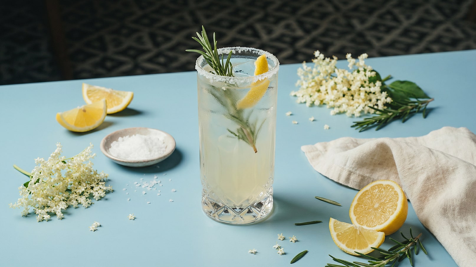 Ein erfrischendes Getränk in einem hochglänzenden Glas mit einem Zuckerrand, garniert mit einem Zitronenstück und einem Rosmarinzweigen. Um das Glas herum liegen frische Zitronenscheiben, Salz und weiße Blüten auf einem blauen Tisch.