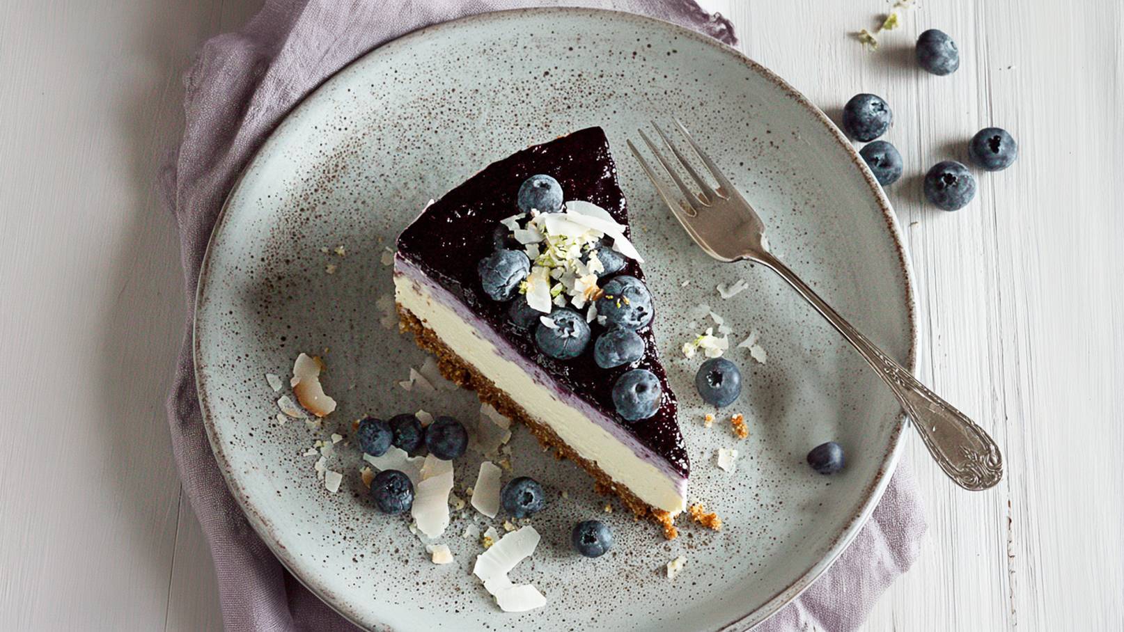 Ein Stück Käsekuchen auf einem grauen Teller, garniert mit blauen Beeren und weißen Kokosraspeln. Eine Gabel liegt neben dem Kuchen, und einige Beeren sind auf dem Teller verteilt.