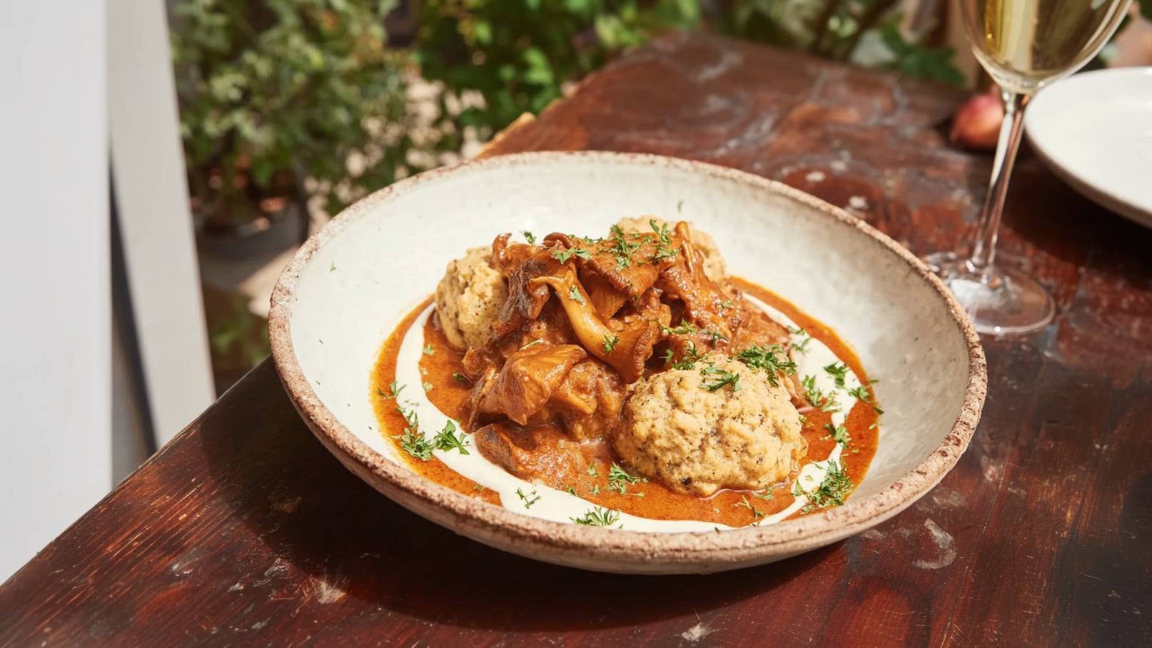 Ein Teller mit einer herzhaften Pilzragout, umgeben von zwei knusprigen Klößen, serviert auf einer weißen Soße. Das Gericht ist mit frischen Kräutern garniert und steht auf einem hölzernen Tisch, im Hintergrund sind unscharfe Pflanzen sichtbar.