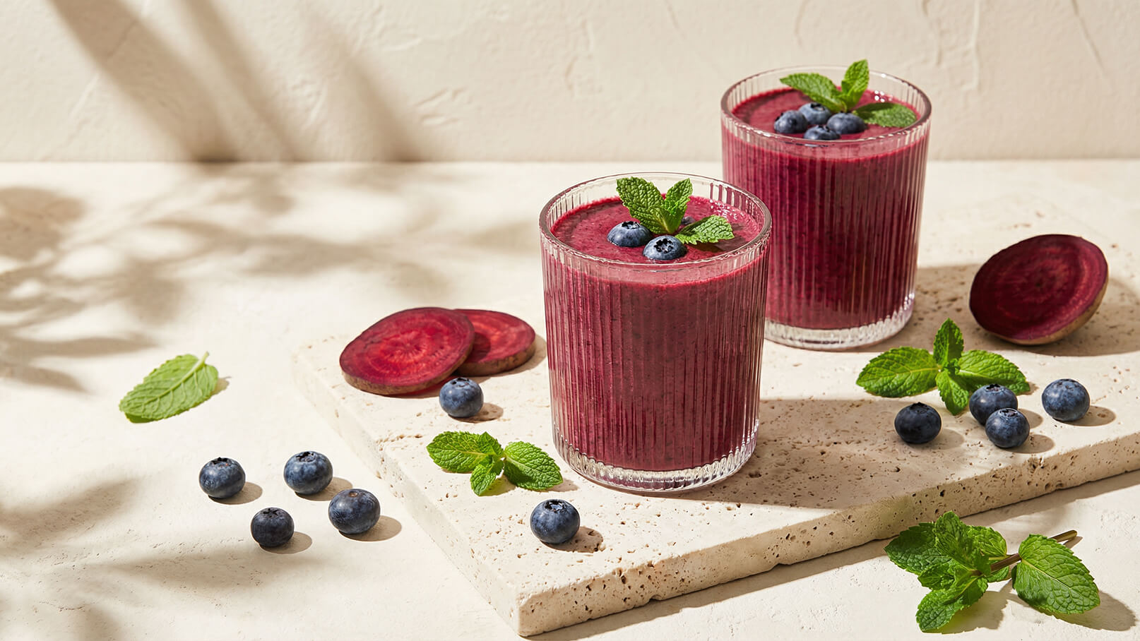 Zwei Gläser mit lila Smoothie stehen auf einem hellen Untergrund. Die Gläser sind mit frischen Blaubeeren und Minzblättern garniert. Neben den Gläsern liegen einige Blaubeeren und eine Scheibe Rote Bete.