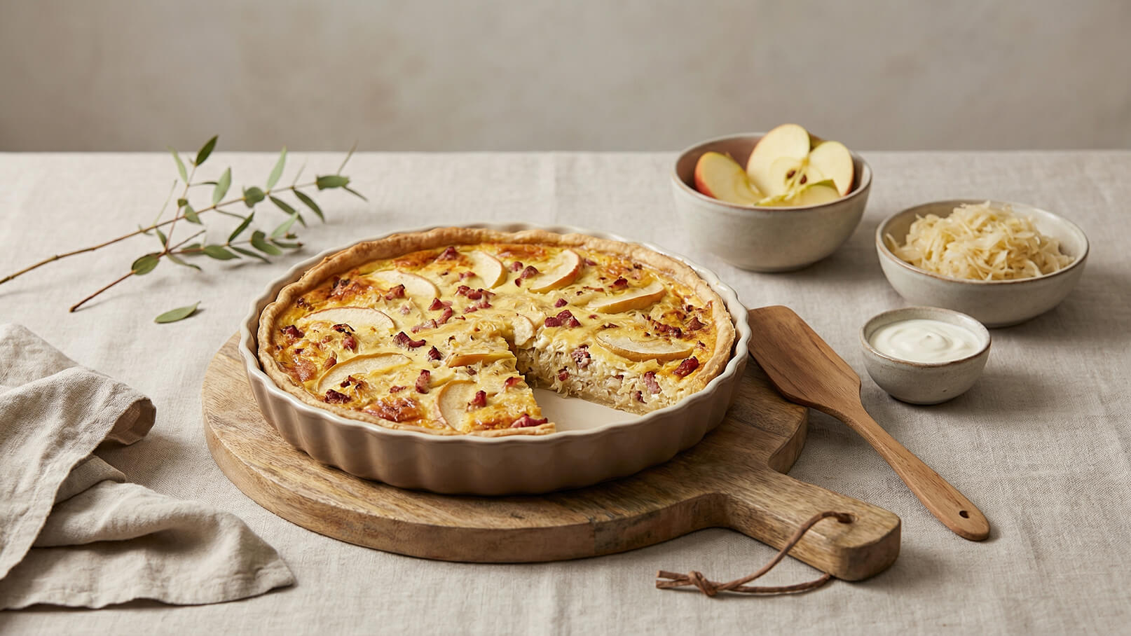 Ein herzhaftes Quichegericht mit einer goldbraunen Kruste, belegt mit Apfelscheiben und Stückchen von Speck. Ein Stück der Quiche wurde herausgenommen. Daneben liegen frische Äpfel, Sauerkraut und eine kleine Schüssel mit einer weißen Sauce auf einem Holzteller.