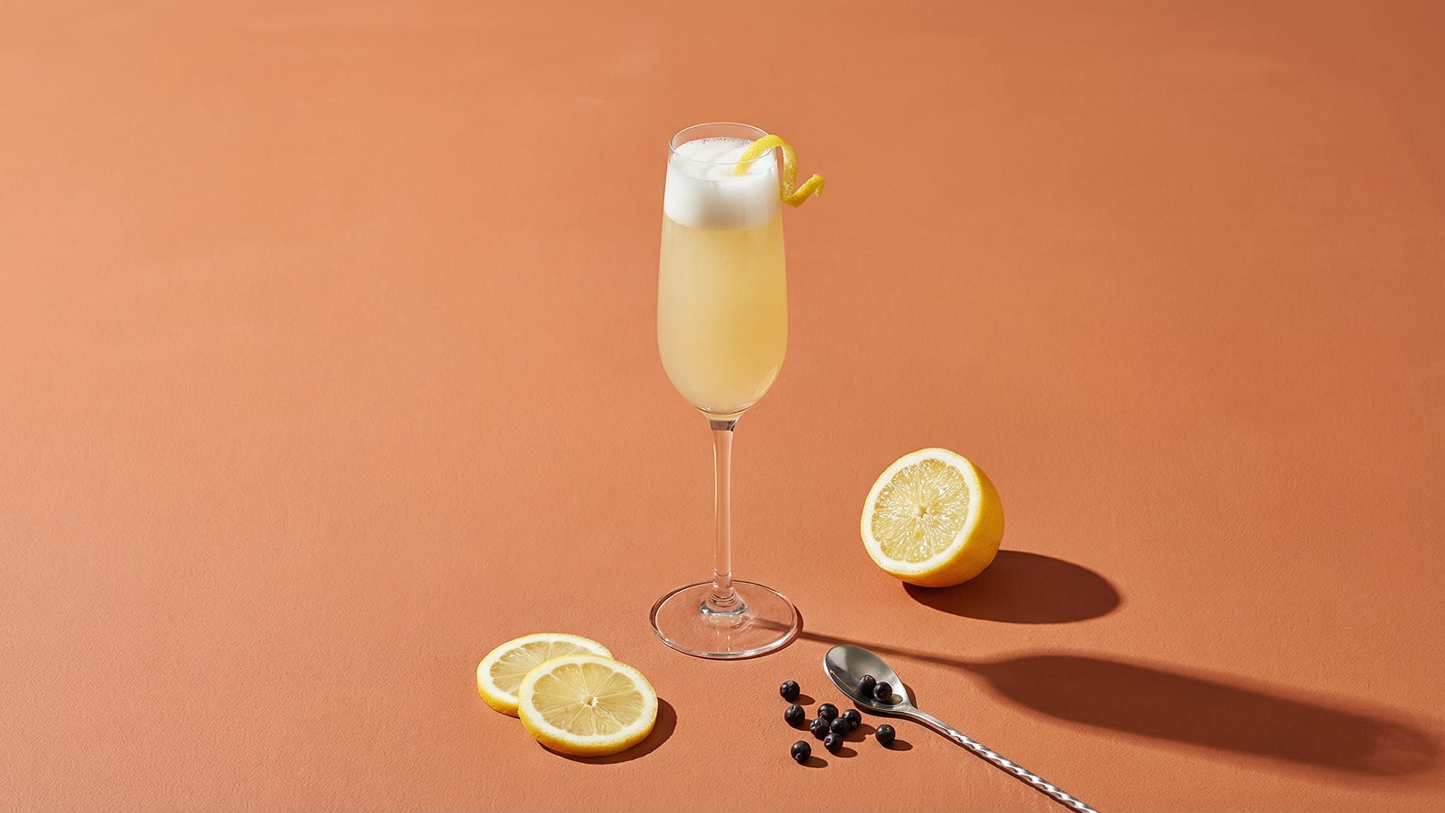 Ein elegantes Glas mit einem schaumigen, gelben Getränk steht auf einem orangefarbenen Untergrund. Neben dem Glas liegen zwei dünne Zitronenscheiben, eine halbierte Zitrone und ein Löffel. Die Szene vermittelt ein frisches und erfrischendes Gefühl.