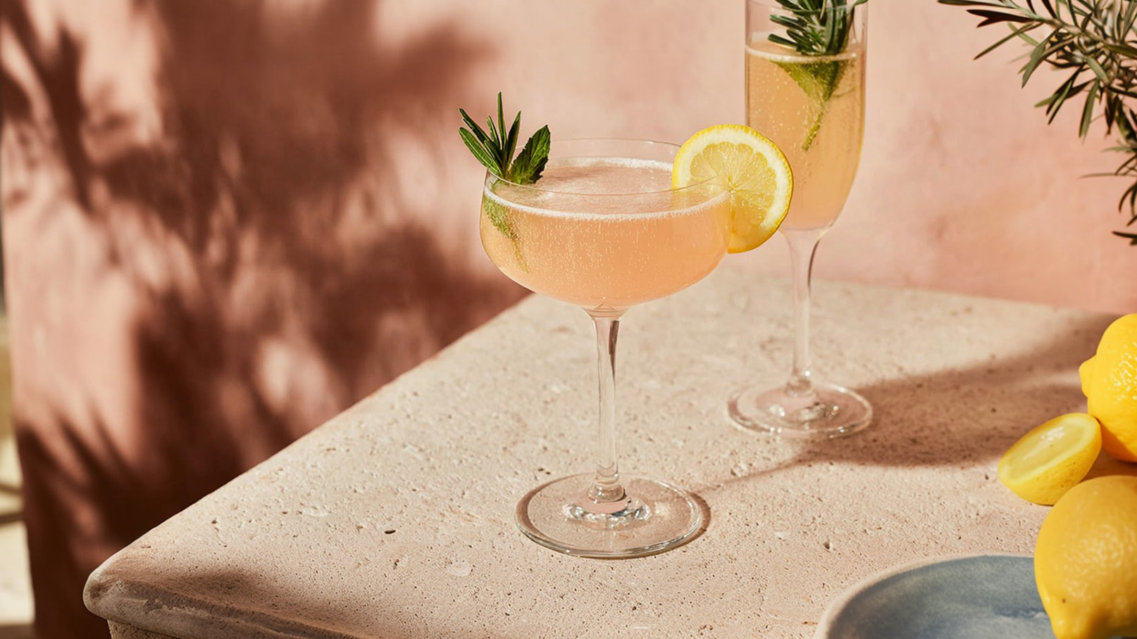 Zwei Cocktails stehen auf einem hellen Tisch. Der erste ist ein Sektglas mit einem Rosmarinzweig, der zweite ein Schaumweinglas mit einem Stück Zitrone und einer Limette. Neben den Gläsern liegen frische Zitronen und ein blauer Teller. Der Hintergrund ist in sanften Rosa-Tönen.