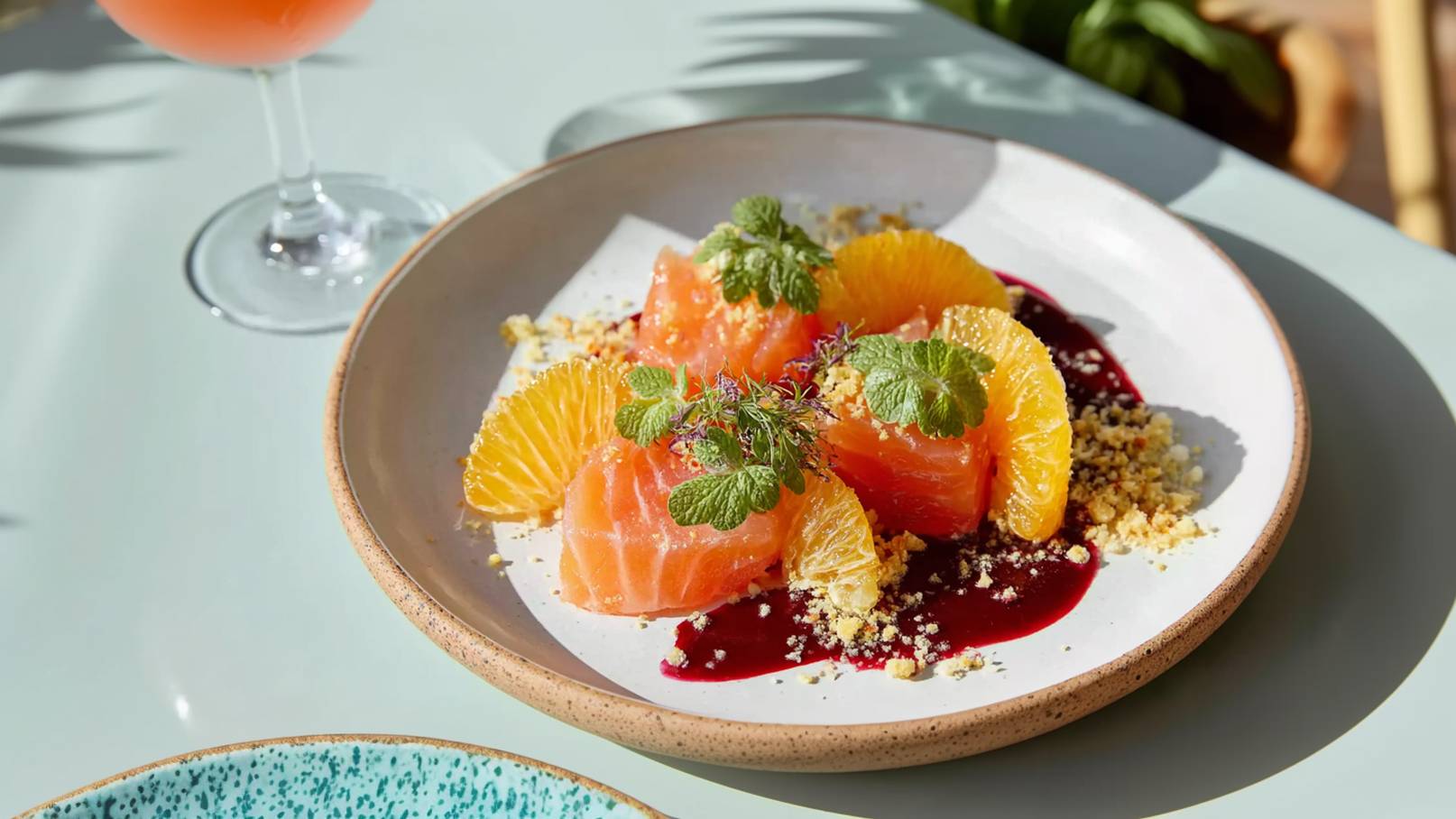 Ein Teller mit Lachsfilets, umgeben von orangefarbener Frucht, garniert mit frischen Kräutern und Bröseln. Der Lachs liegt auf einem fruchtigen Püree in einer weißen Schale mit einem leichten, erdigen Rand. Im Hintergrund ist ein Glas mit einem rosafarbenen Getränk zu sehen.