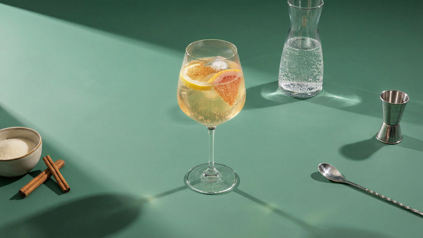 Ein Glaskelch mit einem sprudelnden Getränk, garniert mit Zitronen- und Grapefruitscheiben, steht auf einem grünen Tisch. Daneben befindet sich eine Glaskaraffe mit Wasser und ein Messbecher aus Metall. Im Vordergrund liegt eine kleine Schale mit Zucker und Zimtstangen.