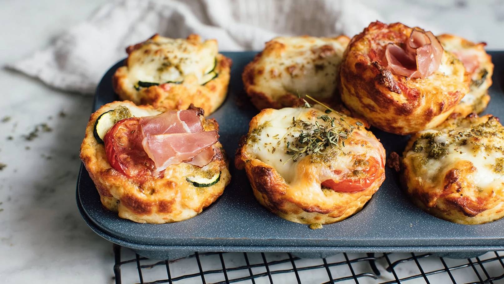Ein Blech mit kleinen, herzhaften Gebäckstücken, gefüllt mit Tomaten, Zucchini und Käse. Einige sind mit Schinken belegt, andere mit Kräutern bestreut. Die Oberflächen sind goldbraun und knusprig, die Stücke sind warm und frisch gebacken.