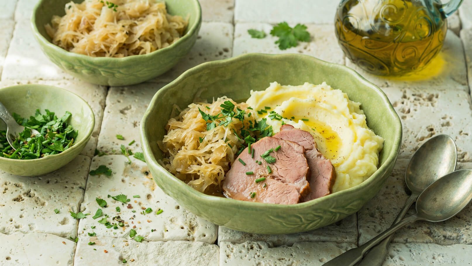 Eine grüne Schüssel enthält ein herzhaftes Gericht mit gekochtem Fleisch, sauer eingelegtem Gemüse und cremigem Kartoffelpüree. Frische Kräuter garnieren das Essen, das auf einem rustikalen Tisch serviert wird. Neben der Schüssel liegen zwei Löffel und eine kleine Schüssel mit gehackten Kräutern.