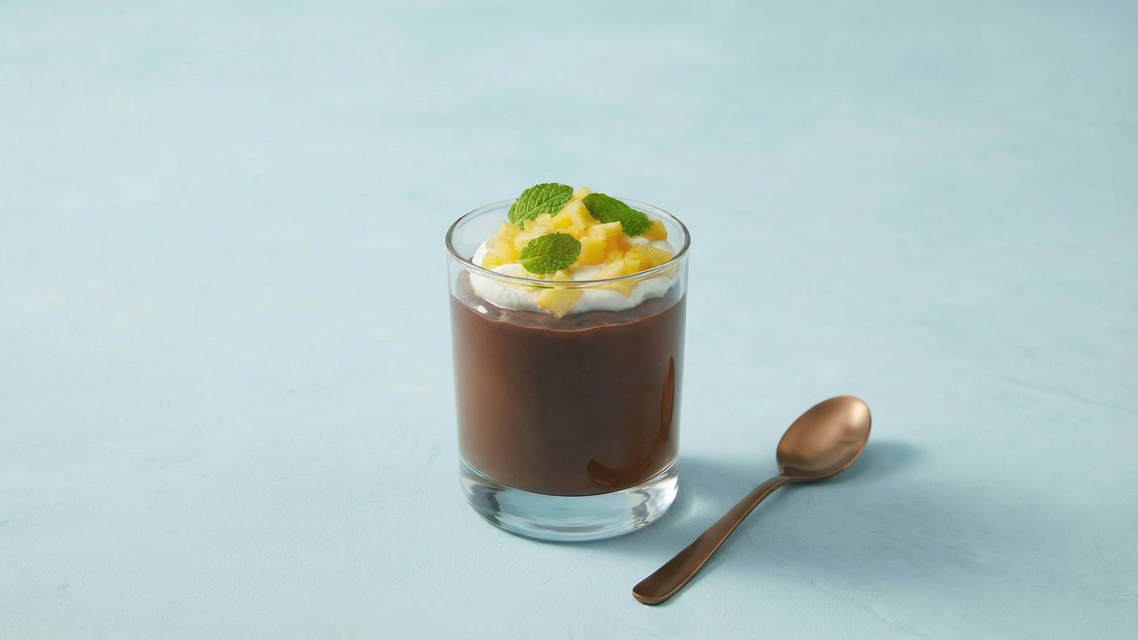 Ein Glas mit Schokoladenpudding, dekoriert mit Schlagsahne, frischen Ananasstücken und Minzblättern. Daneben liegt ein brauner Löffel. Der Hintergrund ist hellblau.