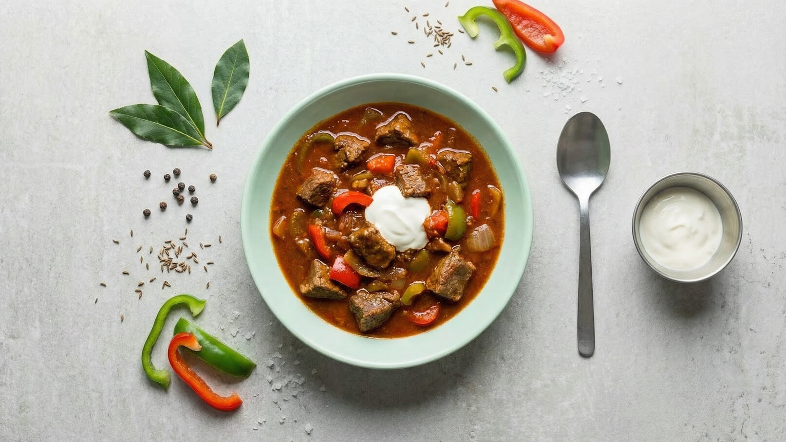 Eine grüne Schüssel mit einem Eintopf aus Fleischstücken und buntem Gemüse wie Paprika und Zwiebeln. Oben ist ein Klecks Joghurt. Neben der Schüssel liegen Gewürze und Paprikastücke. Ein Löffel liegt bereit.