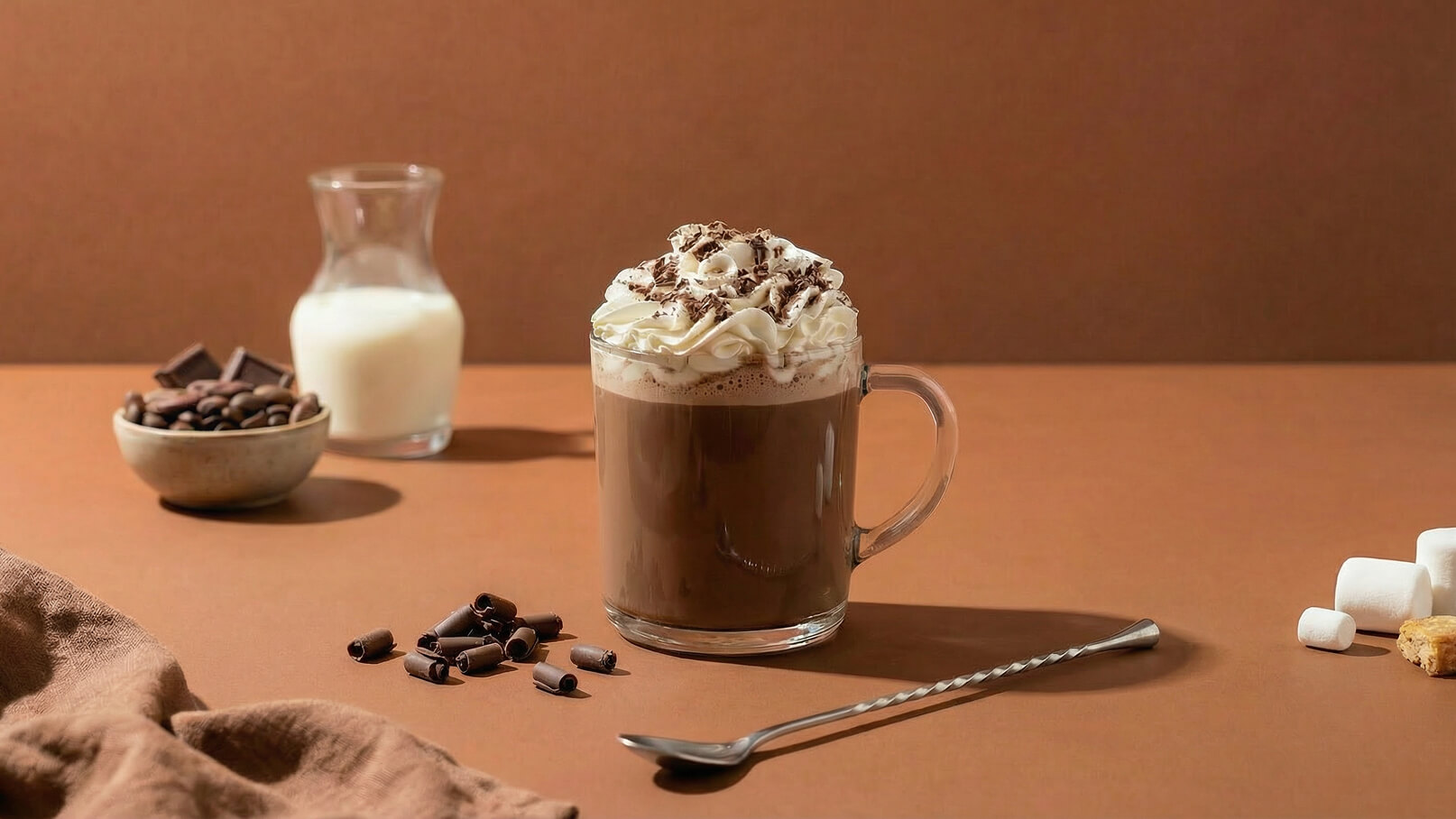 Ein frisch zubereiteter Schokoladengetränk in einem transparenten Glas, dekoriert mit Schlagsahne und Schokoladenstückchen. Daneben liegen Kaffeebohnen und Schokoladenstückchen, sowie ein Glas Milk. Im Vordergrund sind einige Schokoladenröllchen und kleine Marshmallows zu sehen.