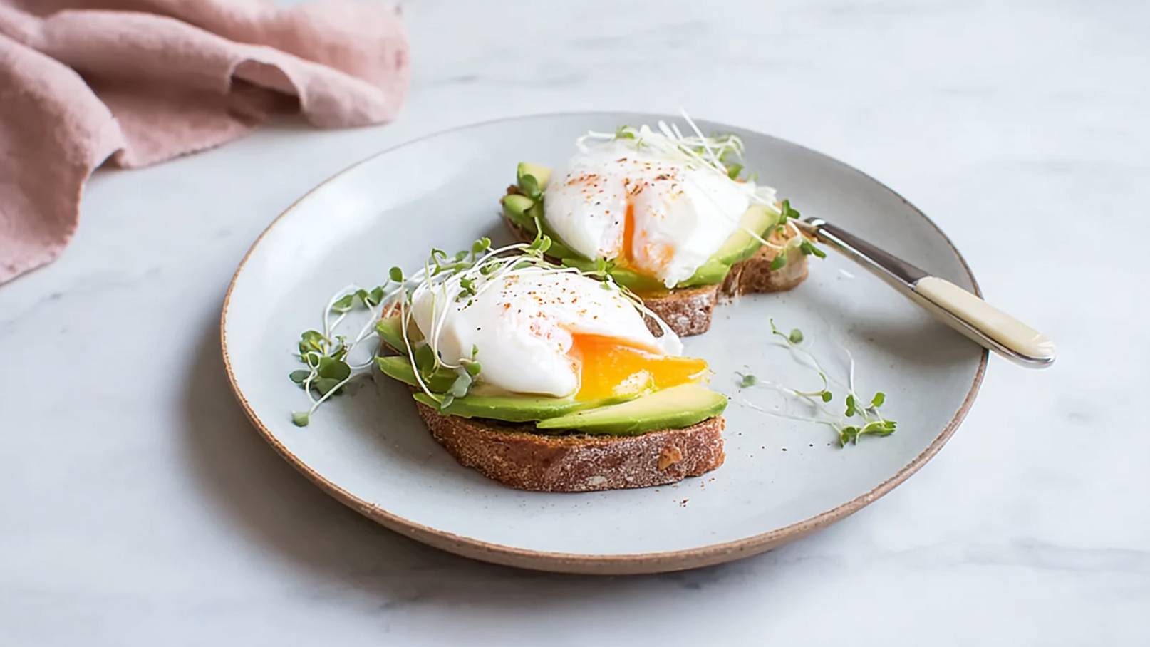 Zwei Scheiben Vollkornbrot belegt mit Avocado und einem pochierten Ei. Das Ei hat einen flüssigen Dotter und ist mit frischen Kräutern garniert. Der Teller steht auf einer hellen Oberfläche, neben einem Besteck.