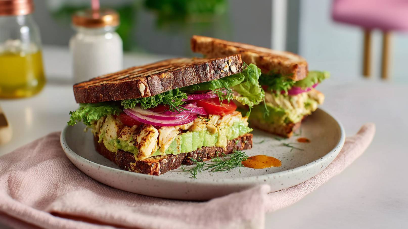 Ein anschaulicher Teller mit einem Sandwich aus Vollkornbrot. Darin befinden sich gegrilltes Hähnchen, Salat, rote Zwiebeln und Tomaten. Das Sandwich ist mit einem leichten Dressing garniert und auf einem rosa Serviette platziert. Im Hintergrund sind eine Flasche Olivenöl und eine Zuckerdose zu sehen.