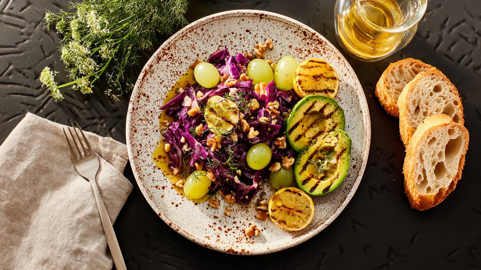 Eine Runde Schale mit lila Krautsalat, grünen Trauben und gehackten Walnüssen. Gegrillte Avocado und Zitrone ergänzen das Gericht. Daneben liegen einige Brotscheiben und ein Glas Öl. Der Hintergrund ist dunkel.