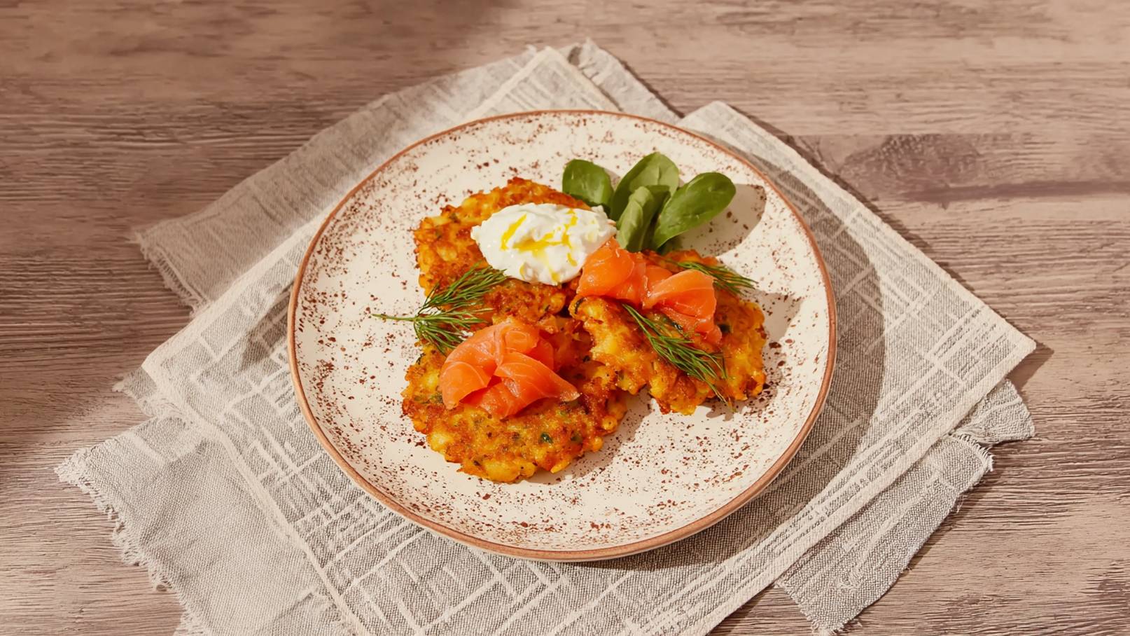 Ein Teller mit knusprigen Kartoffelpuffern, garniert mit Räucherlachs, einem Klecks saurer Sahne und frischen Dillzweigen. Neben dem Puffer liegen einige Blätter frischer Spinat. Der Teller befindet sich auf einem hellen Tisch.
