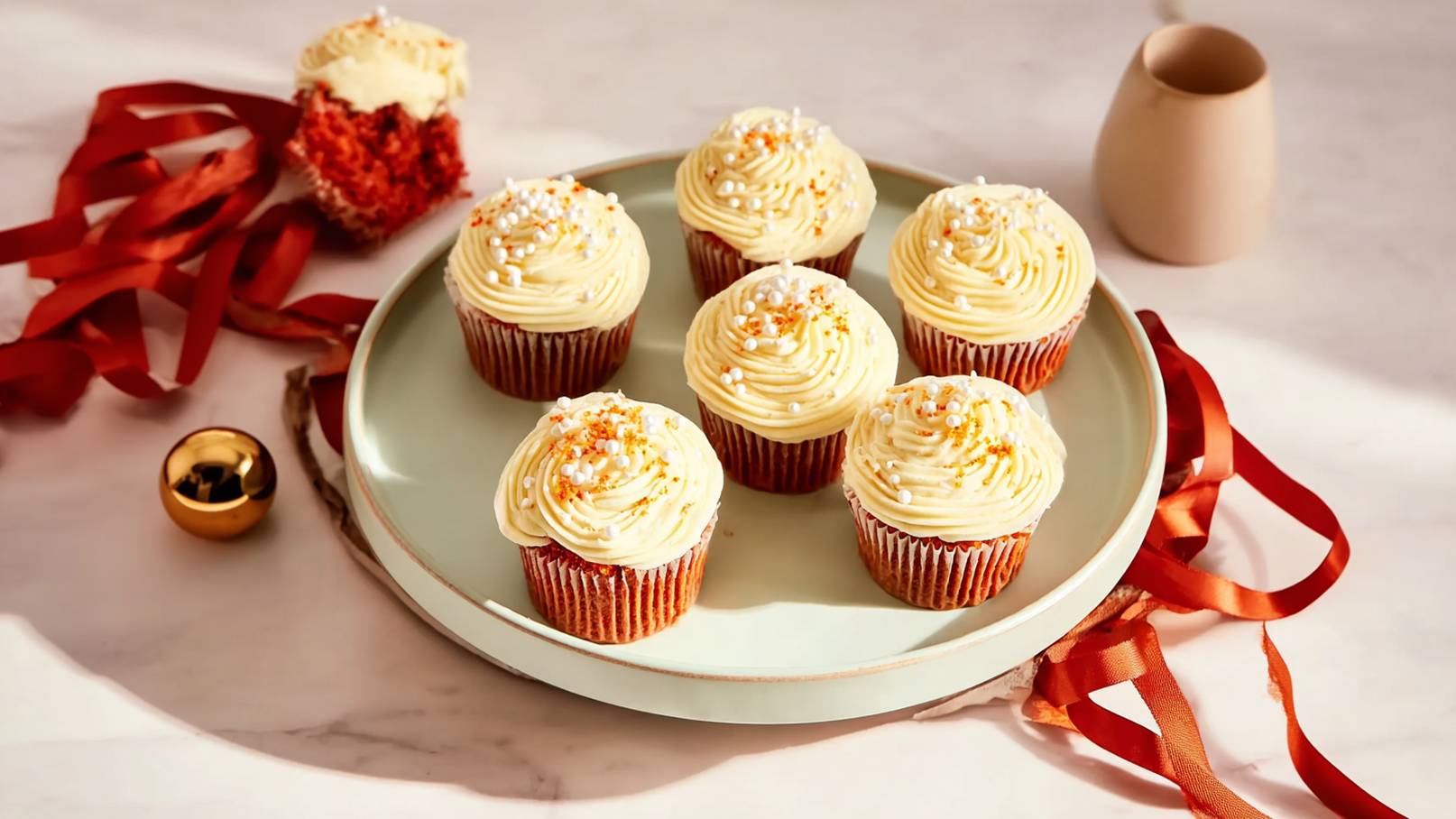 Sechs üppige Cupcakes mit cremigem, hellgelbem Frosting und dekorativen Zuckerkügelchen stehen auf einem flachen, grünen Teller. Im Hintergrund ist ein halb gegessener Cupcake sichtbar. Rote Bänder und eine goldene Kugel ergänzen die festliche Präsentation.