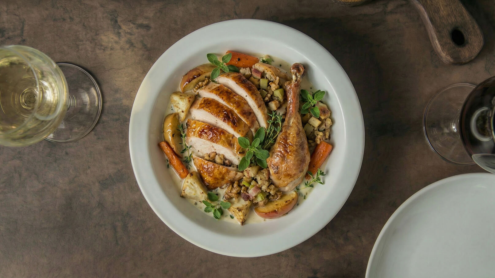 Ein Teller mit gebratenem Hähnchen, das in Scheiben geschnitten ist, liegt auf einem Tisch. Um das Hähnchen herum befinden sich gebratene Karotten, Äpfel und eine Füllung. Frische Kräuter garnieren das Gericht. Im Hintergrund sind ein Glas Weißwein und ein weiteres leeres Geschirr sichtbar.