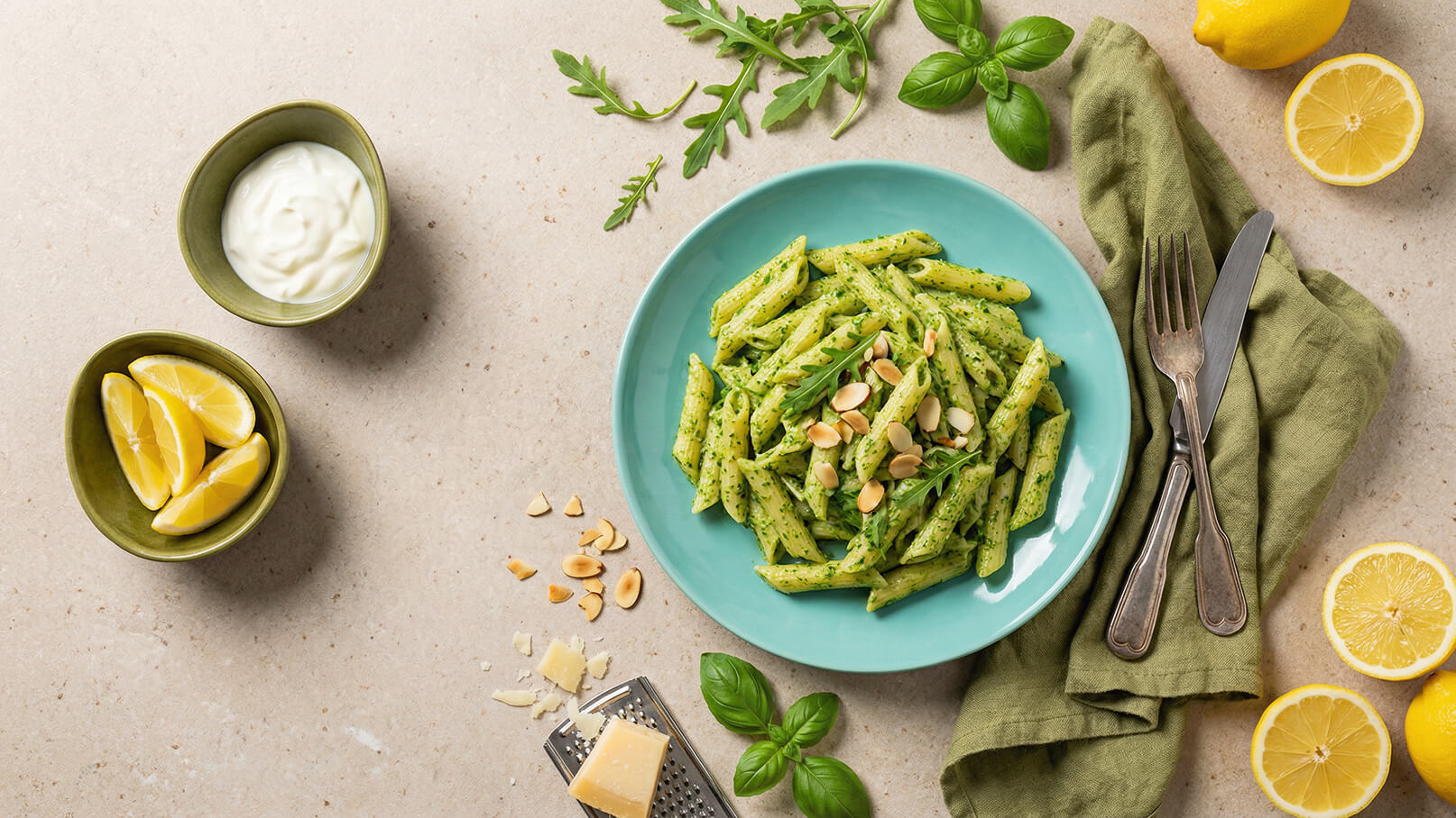 Ein Teller mit grünen Penne-Pasta, garniert mit gehobelten Mandeln und frischem Rucola, steht auf einem türkisfarbenen Tisch. Daneben befinden sich Zitronenschnitze, ein kleiner Schüssel mit Joghurt und frische Basilikumblätter. Ein Messer und eine Gabel liegen auf einem grünen Tuch.