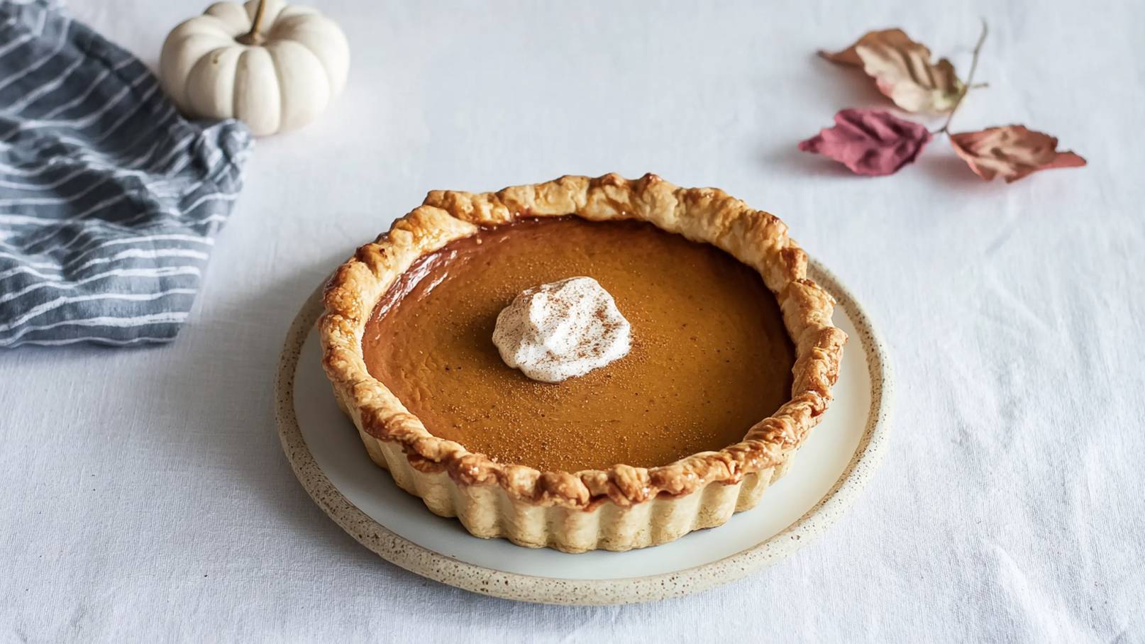 Ein frisch gebackener Kürbiskuchen mit einem goldbraunen Rand und einer cremigen Füllung. Oben prangt ein Klecks Schlagsahne, verfeinert mit Zimt. Der Kuchen steht auf einem hellen Teller, umgeben von einer gestreiften Serviette und einigen bunten Blättern.