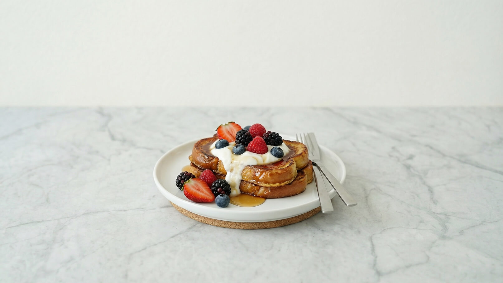 Ein Teller mit zwei goldbraunen Pfannkuchen, die mit Joghurt und einer bunten Mischung aus frischen Beeren, darunter Erdbeeren, Himbeeren, schwarze und blaue Beeren, garniert sind. Ein Schuss Honig läuft über die Pfannkuchen. Gabel und Messer liegen bereit.