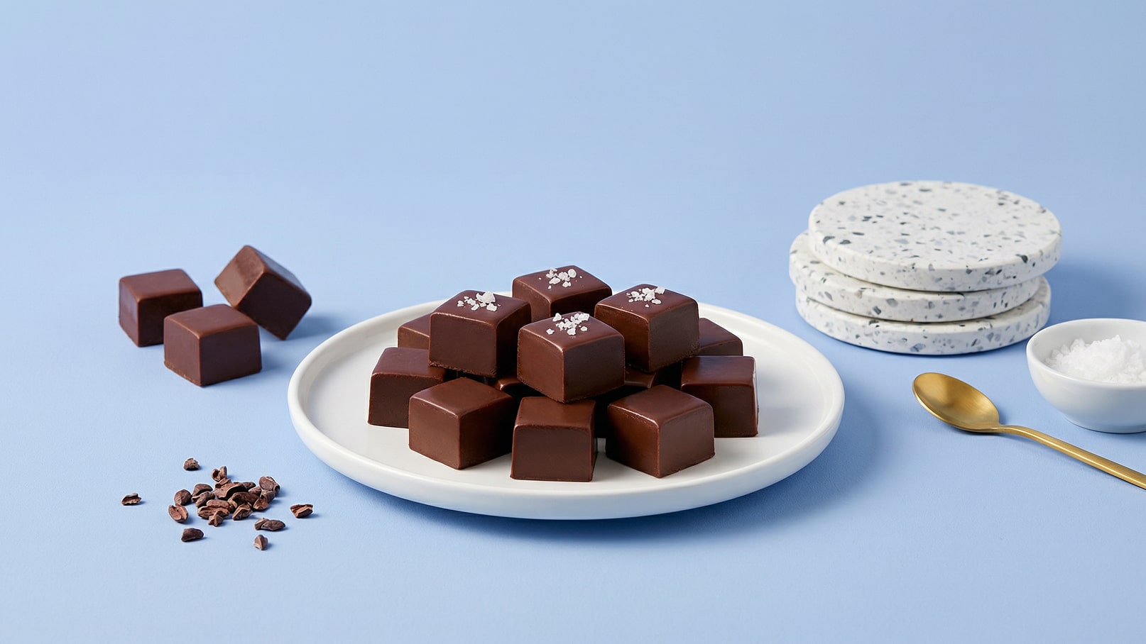 Eine weiße Platte mit einer Anordnung von quadratischen, dunklen Schokoladenstücken, die mit einem Hauch von grobem Salz bestreut sind. Daneben liegen einige Schokoladenstückchen und eine kleine Schüssel mit Salz sowie ein goldener Löffel. Der Hintergrund ist hellblau.