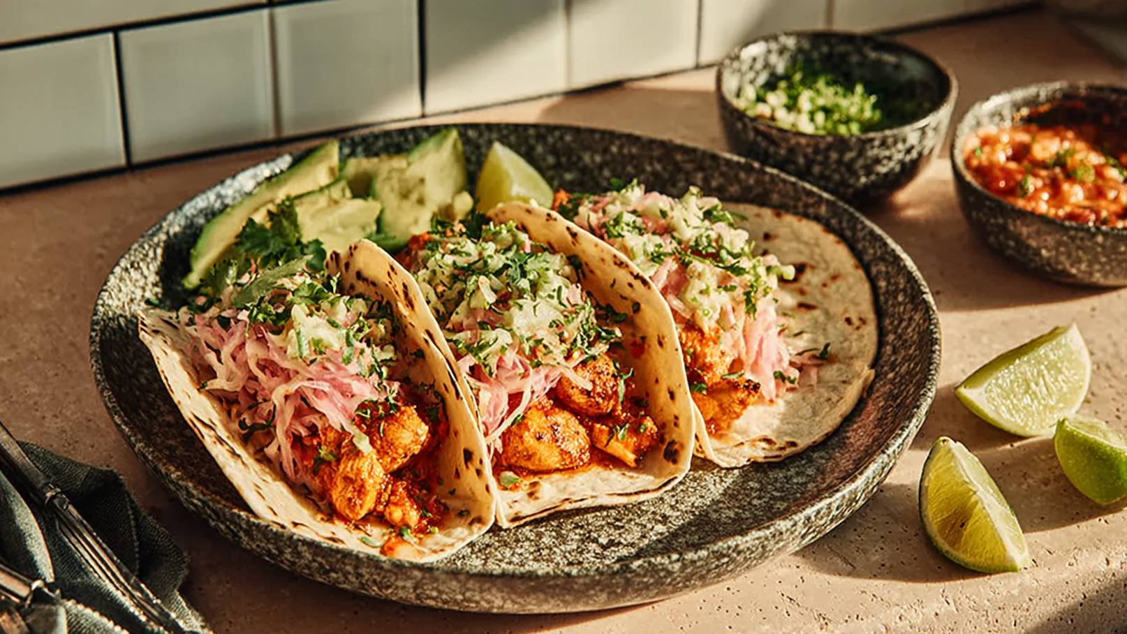 Ein Teller mit drei Tacos, gefüllt mit gegrilltem Fleisch, Weißkraut und frischem Koriander. Daneben liegen Limettenschnitze und guacamole. Das Gericht wird durch kleine Schalen mit Soße und gehobelten Zutaten ergänzt.