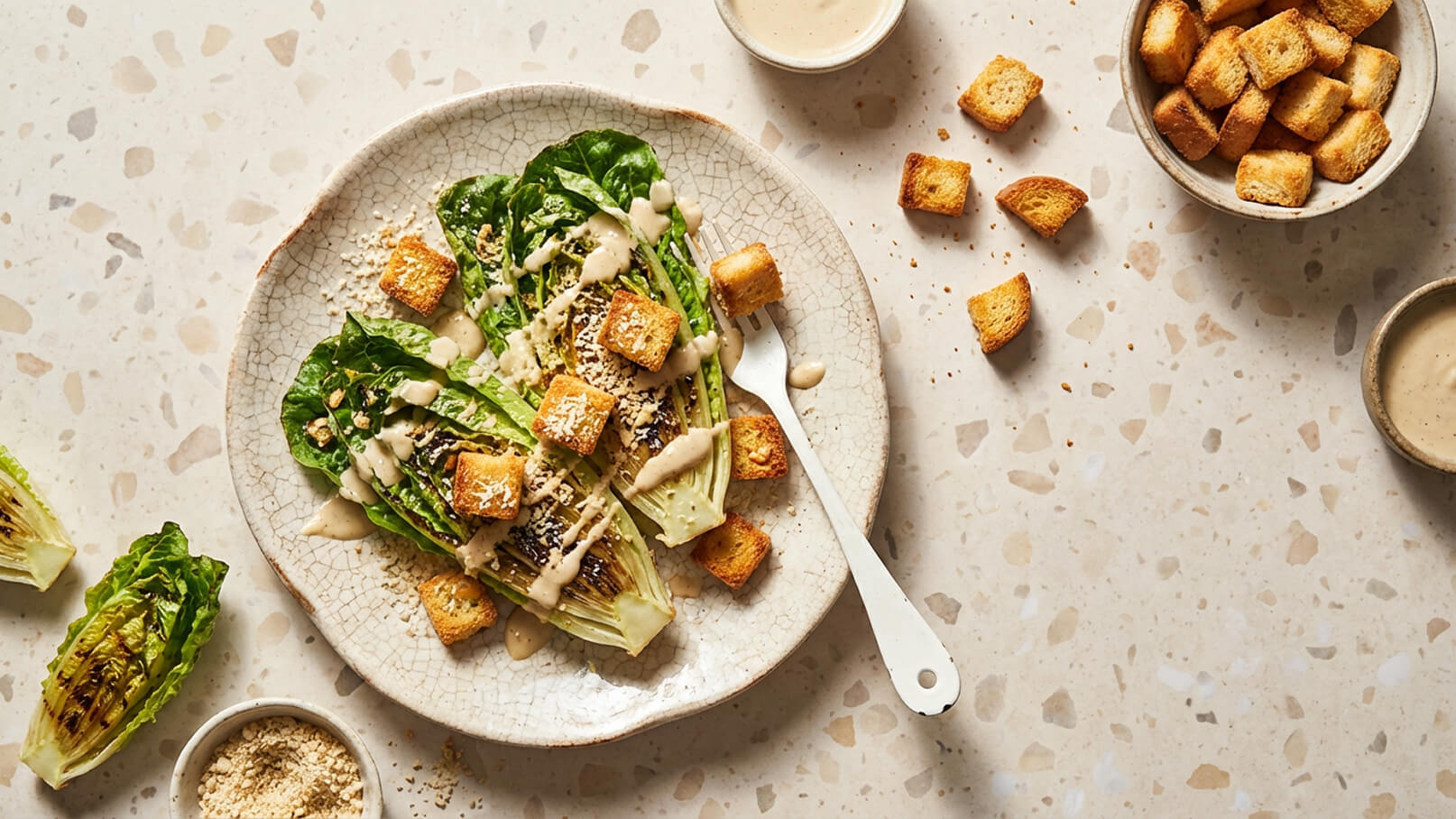 Ein Teller mit gegrilltem Romana-Salat, garniert mit knusprigen Croutons und einem cremigen Dressing. Neben dem Teller liegen weitere Croutons und eine kleine Schale mit Dressing. Der Hintergrund ist strukturiert.