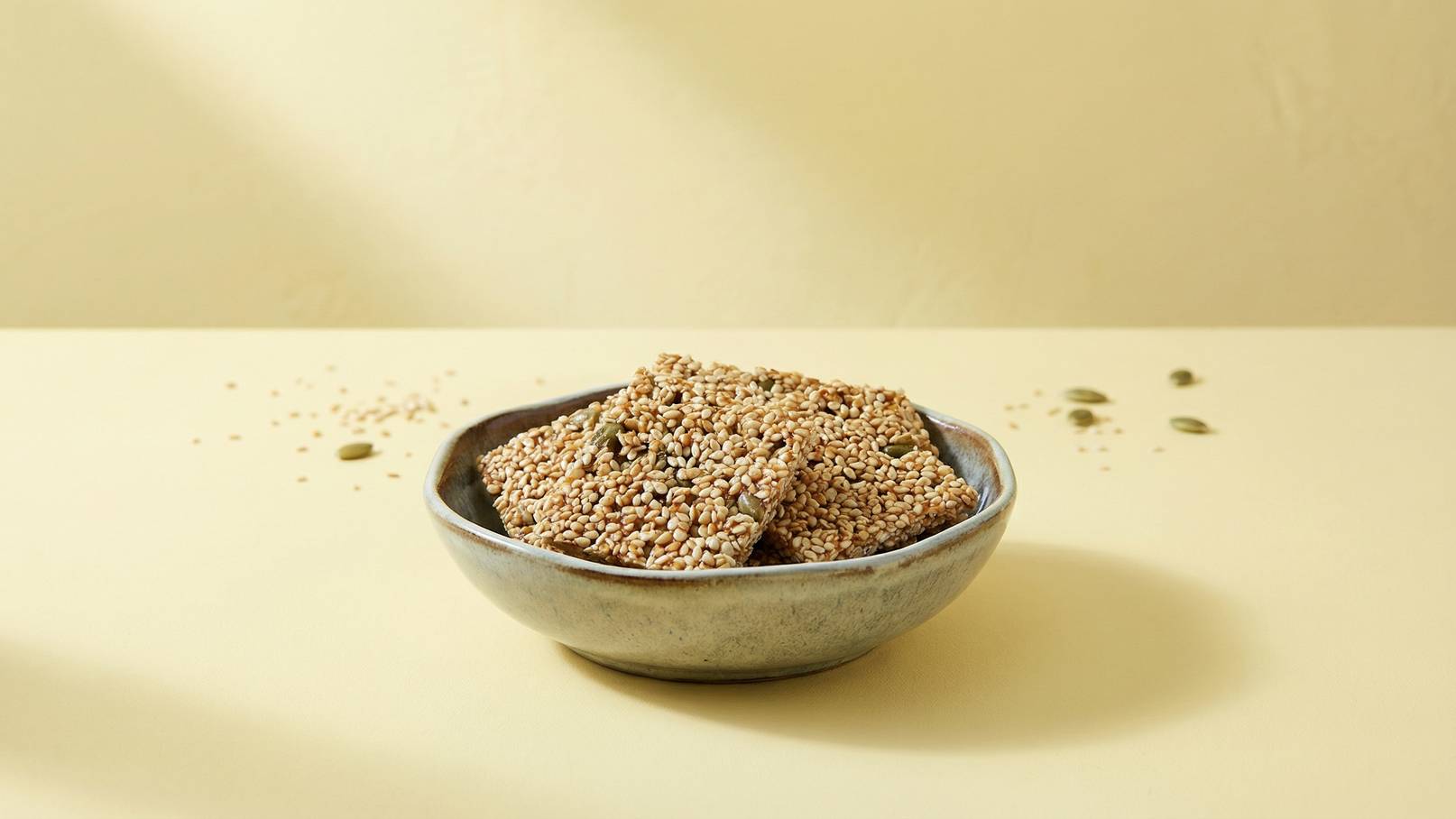Eine kleine Schüssel mit quadratischen, knusprigen Sesamriegeln steht auf einer hellen Oberfläche. Einige zerstreute Sesamsamen sind ringsum sichtbar. Die Riegel sind goldbraun und optisch ansprechend.