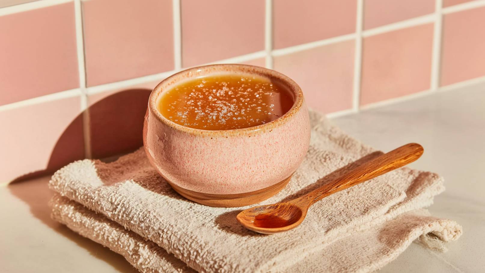 Eine rosa Schüssel mit einer warmen Flüssigkeit steht auf einem Handtuch. Neben der Schüssel liegt ein kleiner Holzlöffel. Die Oberfläche der Flüssigkeit ist leicht schimmernd, und das Gesamtergebnis vermittelt eine gemütliche, einladende Atmosphäre.
