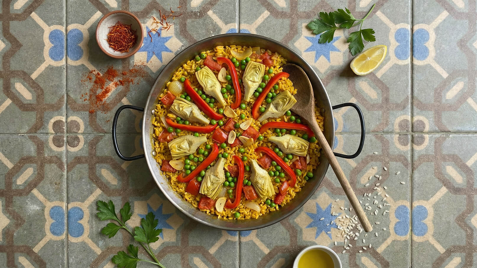 Eine große Pfanne mit Paella, gefüllt mit gelbem Reis, Erbsen, Paprika, Artischocken und Tomaten. Ein Holzlöffel liegt in der Pfanne, und frischer Zitronensaft sowie Petersilie sind daneben platziert. Der Untergrund zeigt farbenfrohe Fliesenmuster.