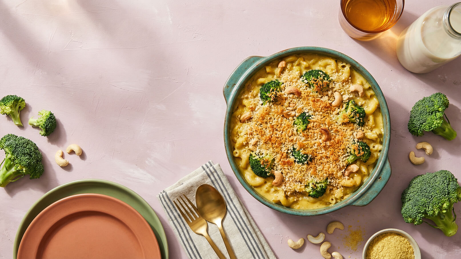 Ein Auflaufgericht in einer blauen Schüssel, überbacken mit einer goldenen Kruste, und verziert mit Brokkoli und Cashewkernen. Daneben liegen zwei Teller, Besteck und eine Streuung von Nahrungsmitteln wie Brokkoli und Cashewkerne auf einer hellen Oberfläche.