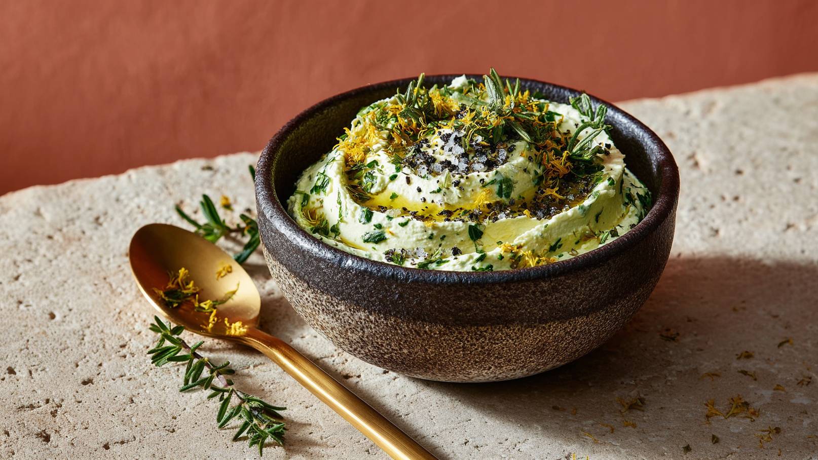 Eine dunkle Schüssel mit cremigem, würzigem Dip ist gefüllt. Der Dip wird mit frischen Kräutern und Gewürzen garniert und hat eine glatte Oberfläche. Daneben liegt ein goldener Löffel, umgeben von einigen Kräuterzweigen.