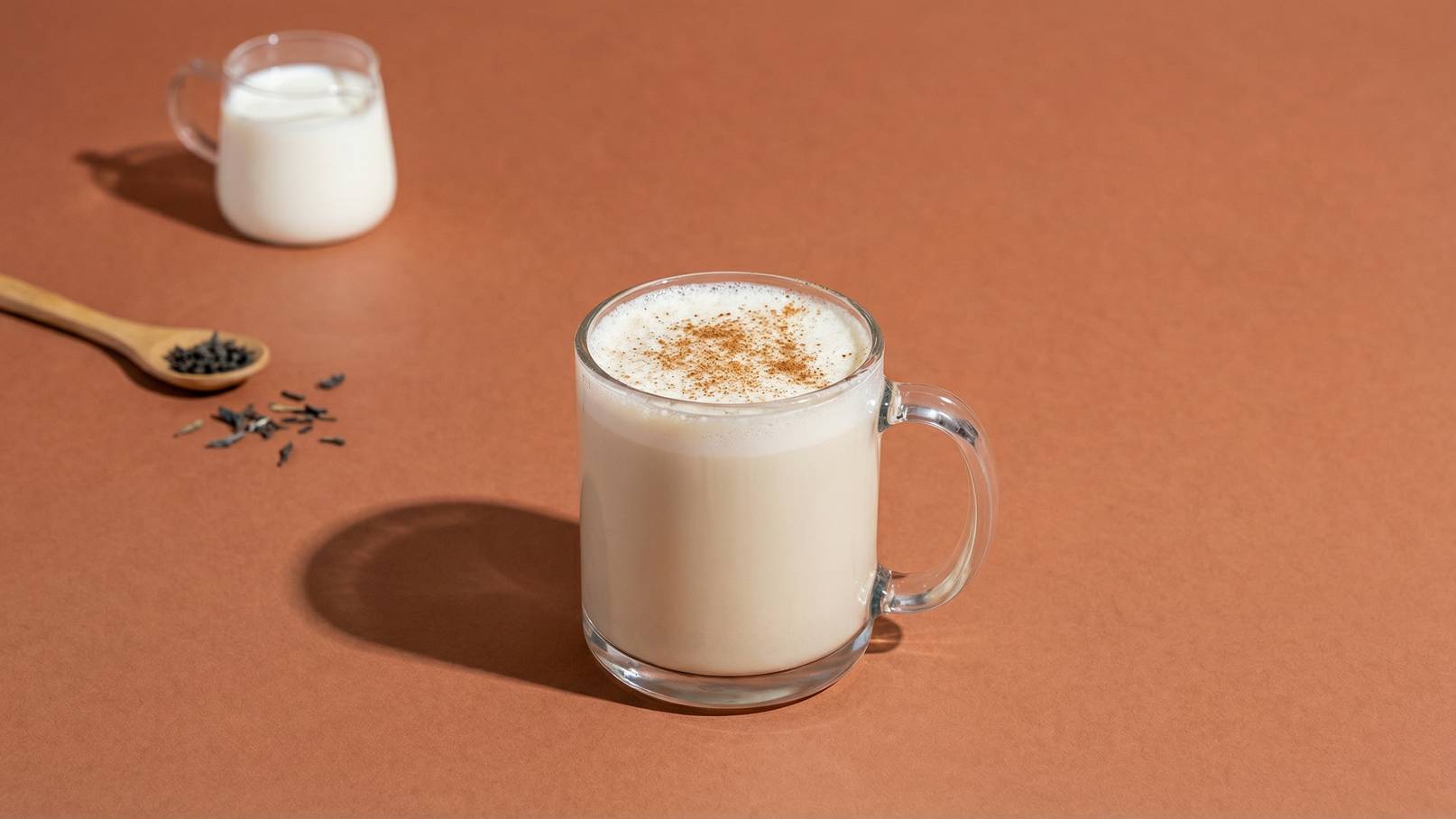 Ein Glas mit cremigem Getränk, bestreut mit Zimt, steht auf einem braunen Untergrund. Im Hintergrund ist ein kleiner Krug mit Milch und ein Löffel mit Gewürzen zu sehen.