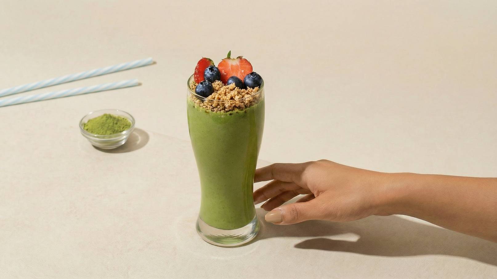 Ein Glas grüner Smoothie ist mit einer Schicht Granola, frischen Erdbeeren und Heidelbeeren garniert. Eine Hand greift nach dem Glas, während zwei gestreifte Trinkhalme und eine kleine Schale mit grünem Pulver im Hintergrund zu sehen sind.