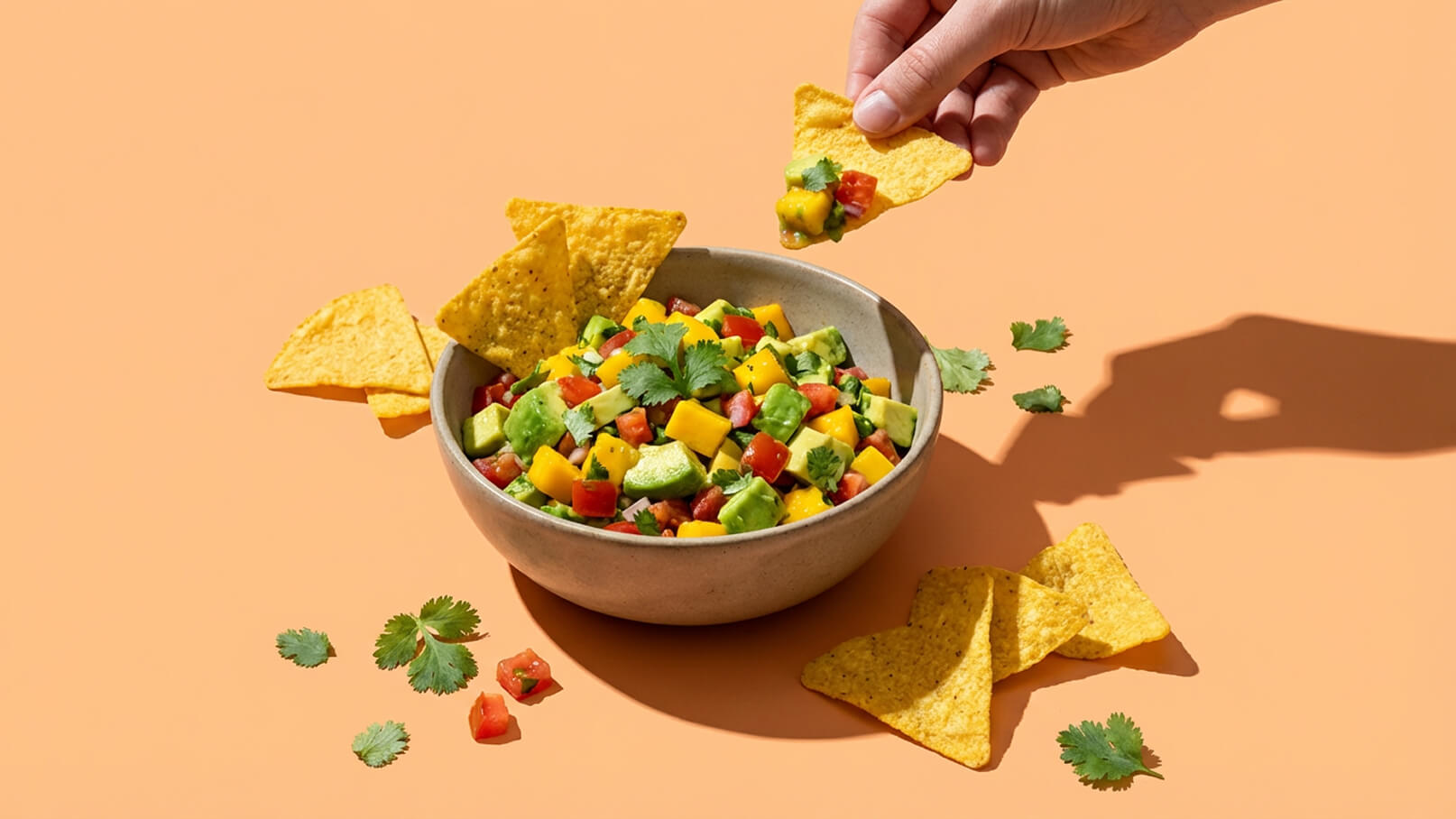 Eine Hand hält eine Tortilla-Chips, die in eine Schüssel mit buntem Salat aus Avocado, Mango, Tomaten und frischem Koriander eintaucht. Um die Schüssel liegen weitere Tortilla-Chips und etwas Koriander. Der Hintergrund ist in einem sanften Orangeton.