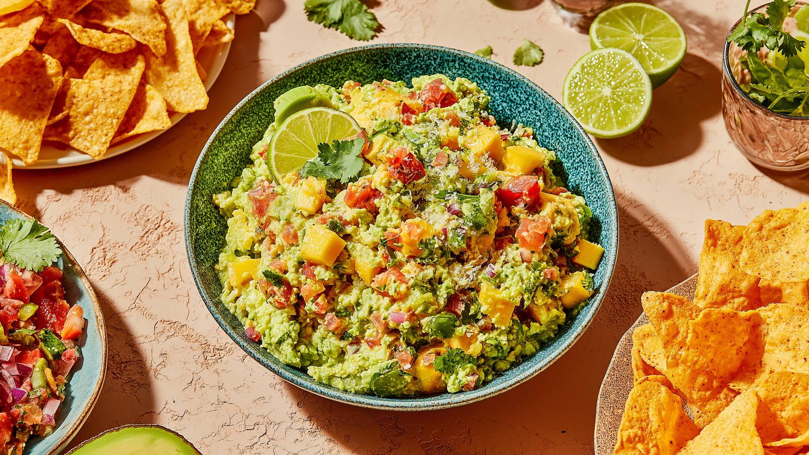 Eine Schüssel mit buntem Guacamole, verfeinert mit Tomaten, Mango und Koriander, ist im Mittelpunkt. Um die Schüssel herum liegen einige Tortilla-Chips auf einem Teller. Frische Limettenscheiben und Kräuter ergänzen das Bild.