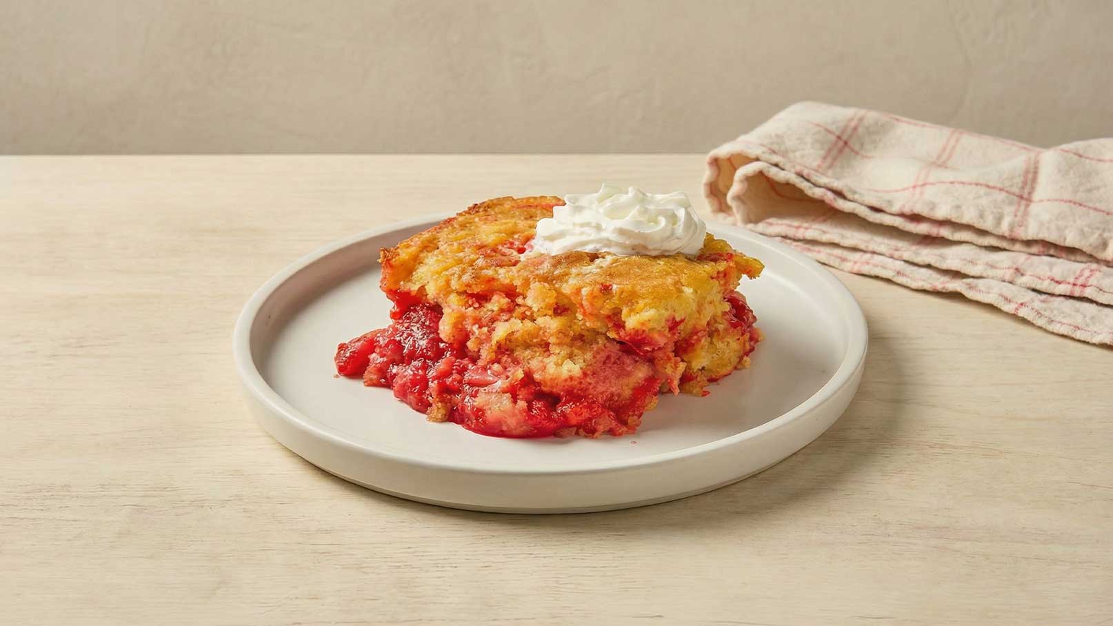 Ein Stück Kuchen auf einem weißen Teller, mit rotem Obstbelag und Sahnehaube. Der Kuchen hat eine goldene, leicht krümelige Textur. Im Hintergrund liegt ein kariertes Geschirrtuch.